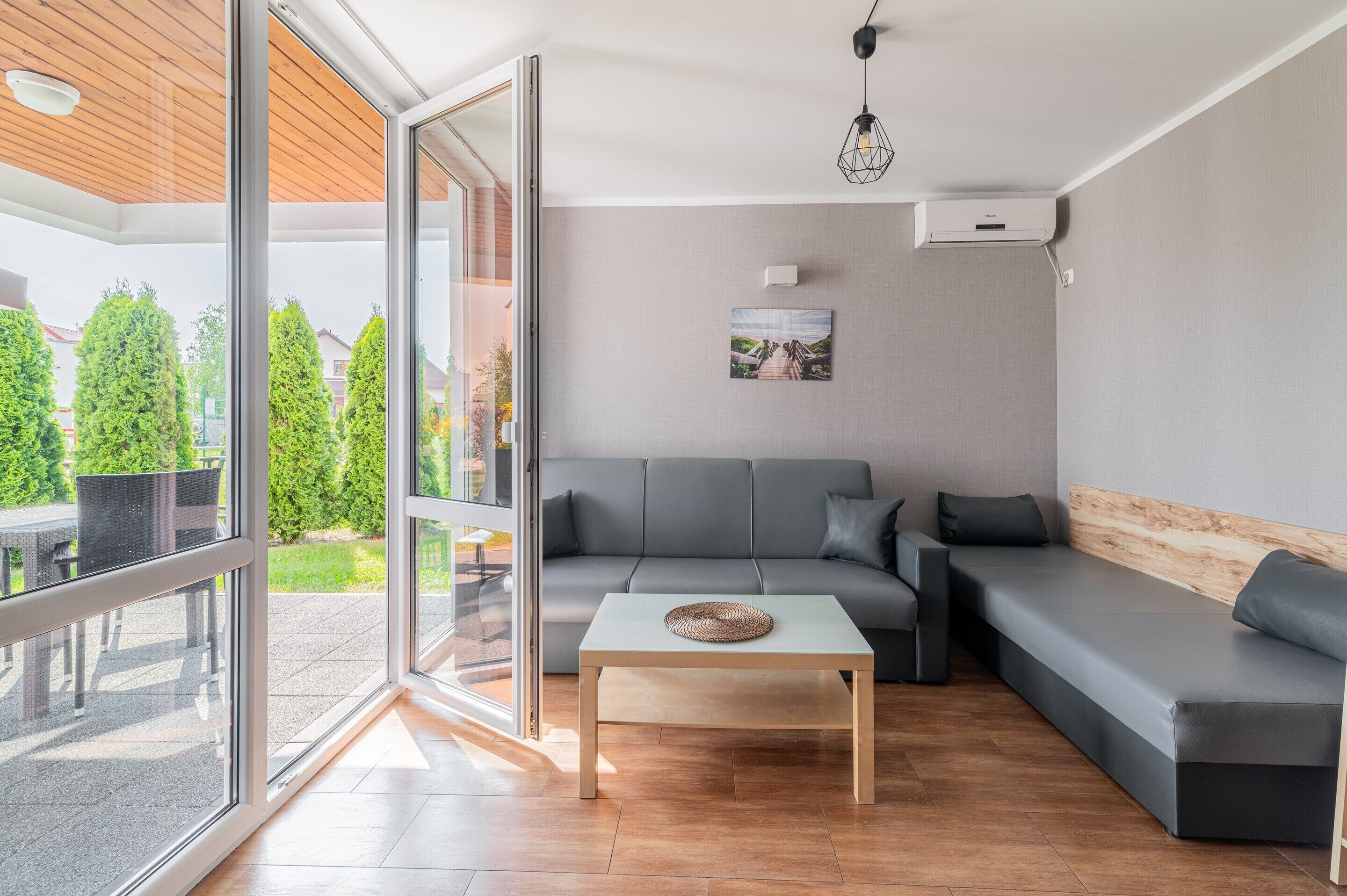Una sala moderna con un sofá gris, una mesa de centro de madera y un ventanal que da a un área exterior con una mesa y sillas de patio.