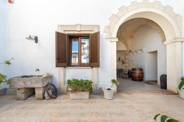 An outdoor area with a stone well, potted plants, a wooden barrel, and an arched doorway leading to a building.