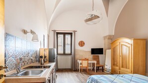 A compact room with a kitchenette, a bed, a wooden wardrobe, a TV, and a window with curtains.