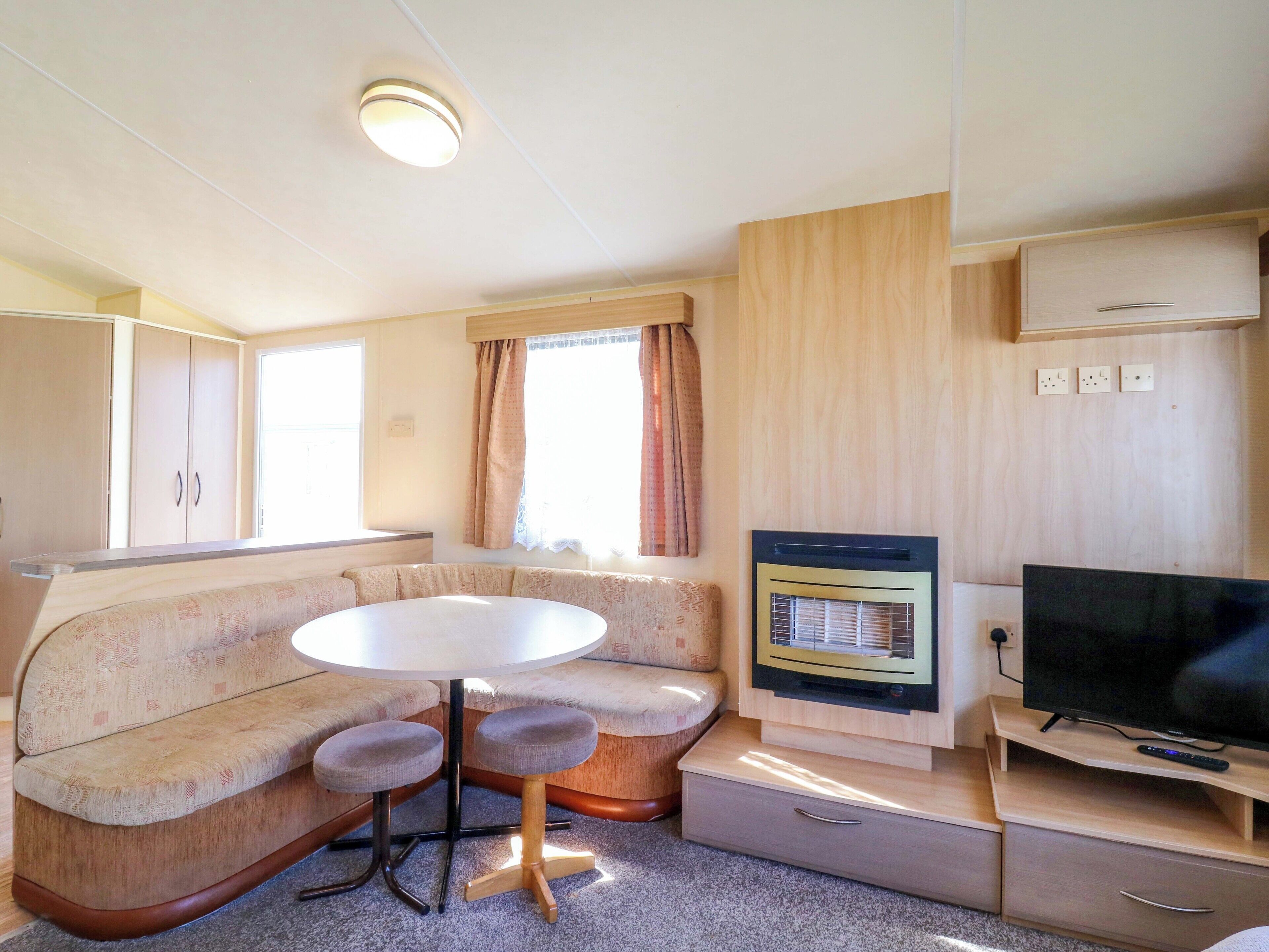 A compact RV interior with a kitchenette, a dining area with a table and two chairs, a television, and a window with curtains.