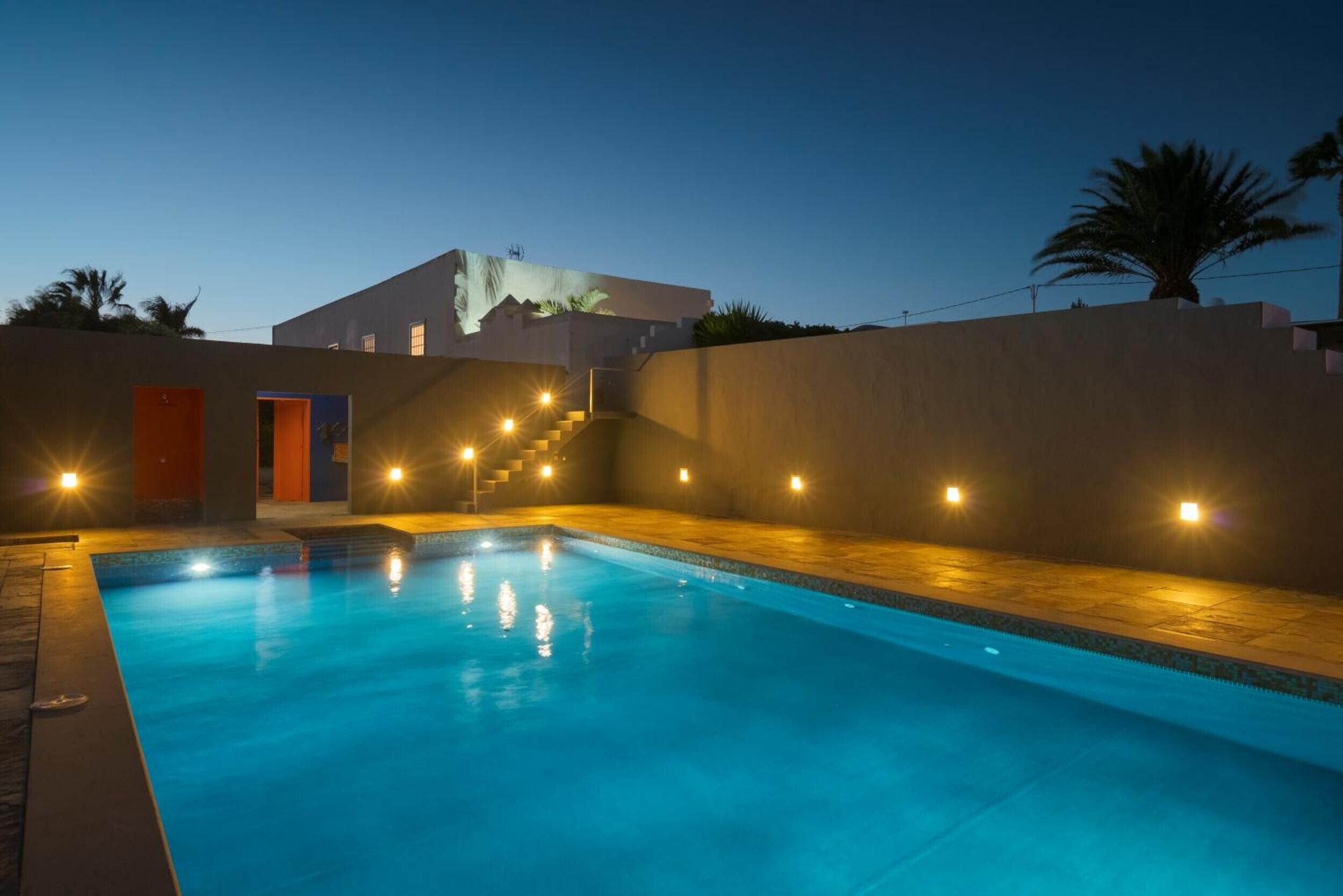 A swimming pool with steps leading to a building, surrounded by walls and a palm tree.