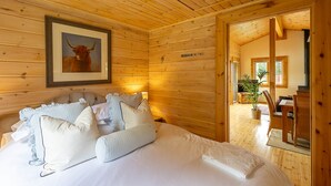 A wooden cabin bedroom with a bed, pillows, a framed picture of a bull, and a dining area visible through an open door.