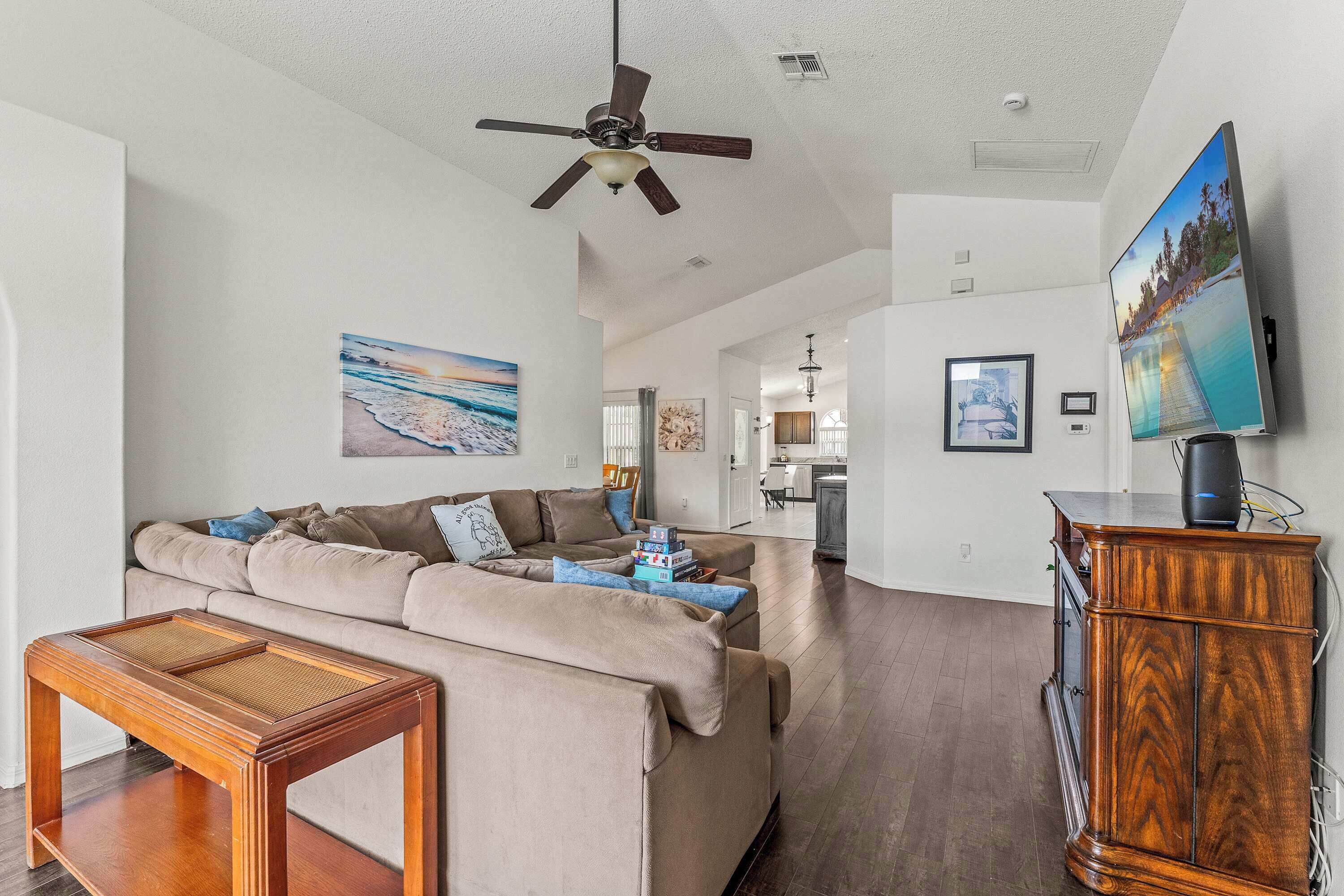 A modern living room with a sectional sofa, a wooden coffee table, a ceiling fan, a flat-screen TV mounted on the wall, and a wooden cabinet.