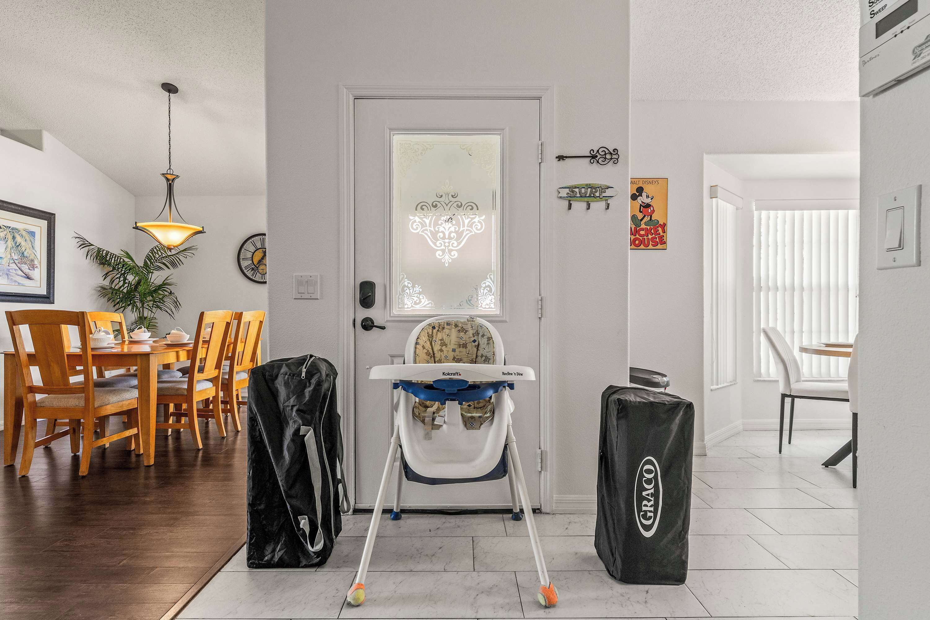 A room with a high ceiling, a dining area with wooden chairs and a table, a baby changing station with a high chair, and a door with decorative glass.