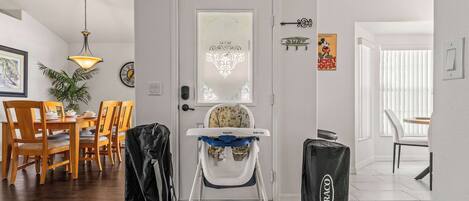 A room with a high ceiling, a dining area with wooden chairs and a table, a baby changing station with a high chair, and a door with decorative glass.