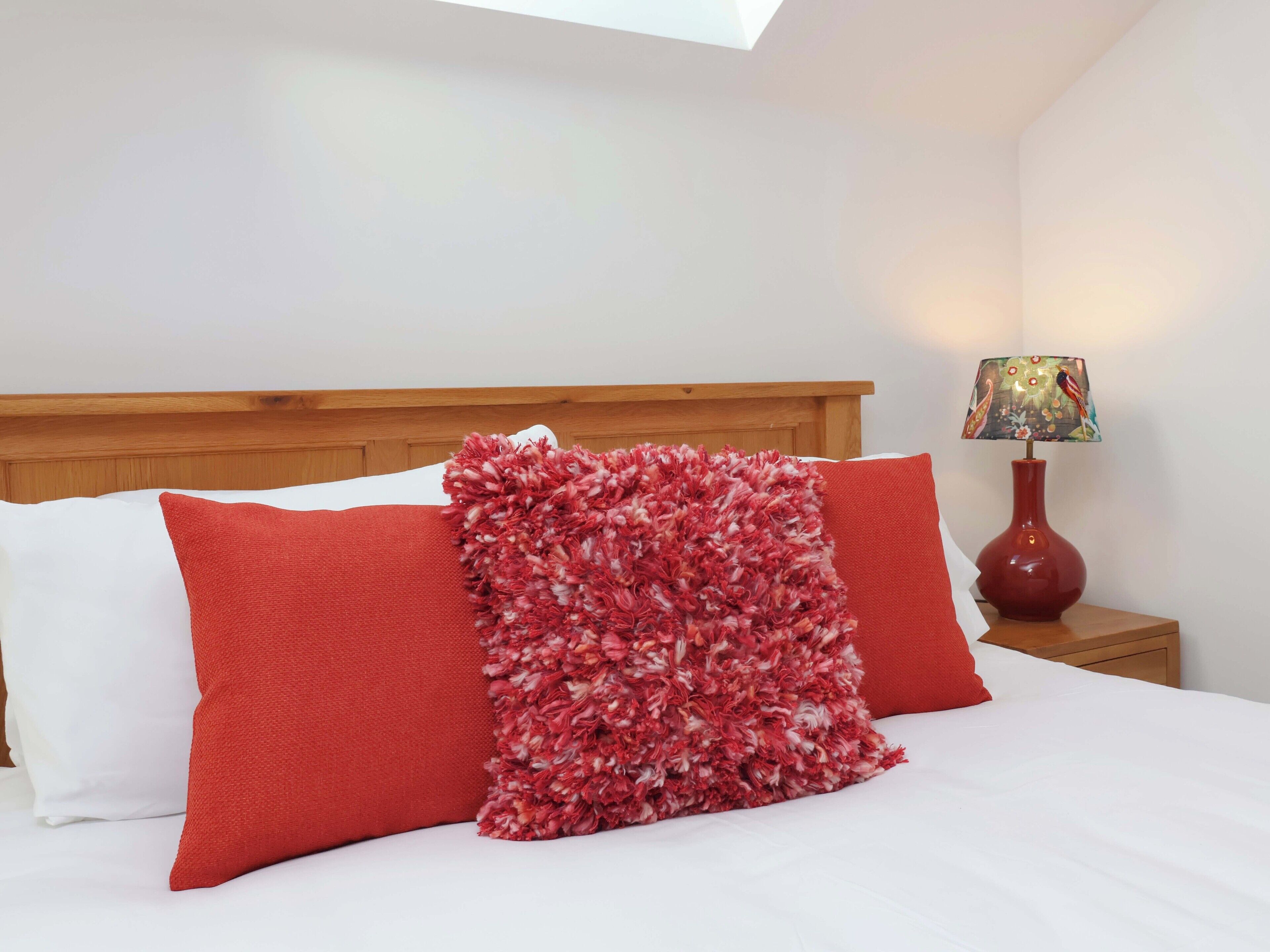 Una cama bien tendida con cabecera de madera, una manta roja y blanca, y un jarrón rojo con una lámpara a su lado.