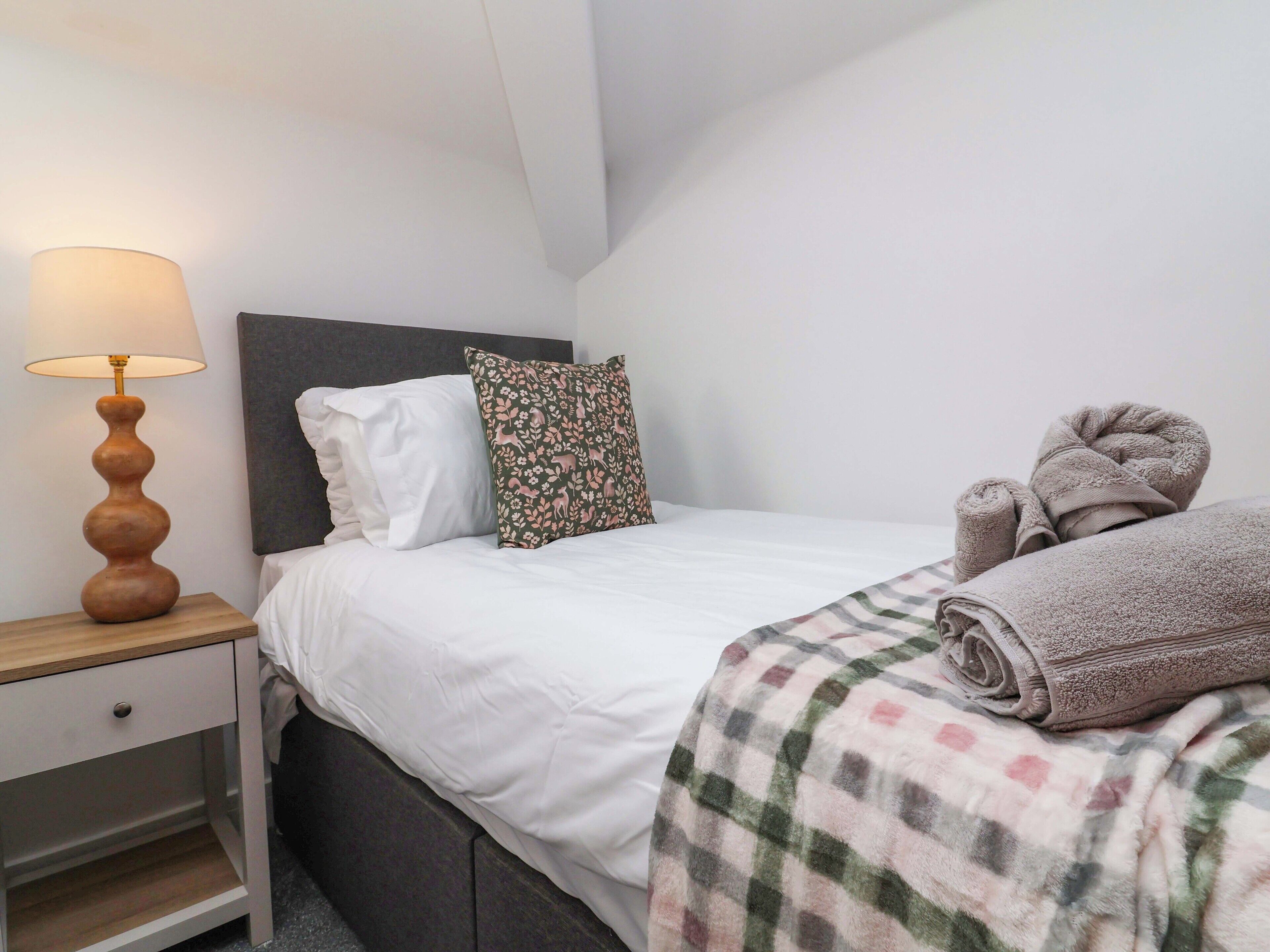 A neatly made bed with white bedding, a patterned pillow, a rolled towel, and a bedside lamp on a wooden nightstand.