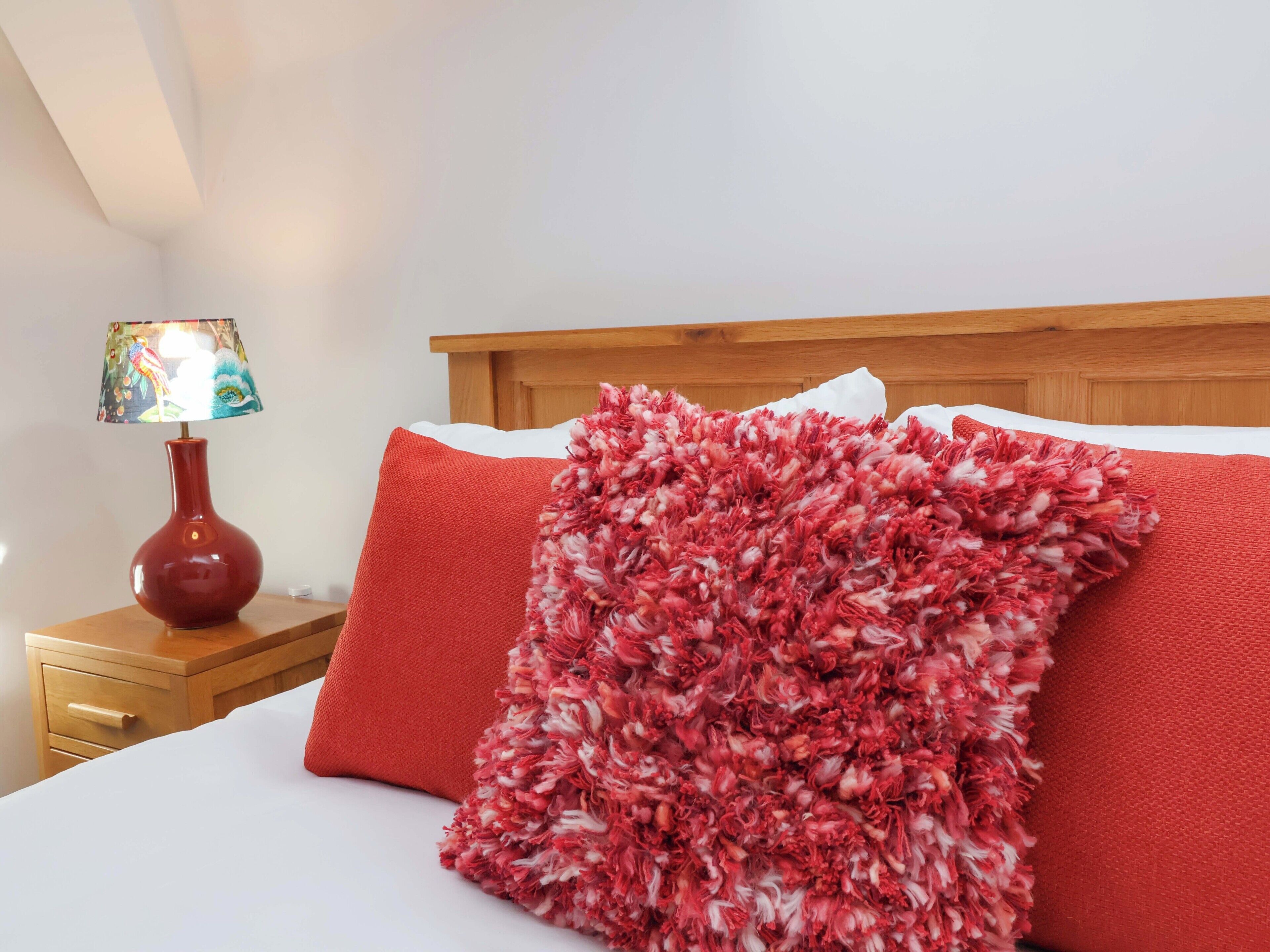 Uma cama com travesseiro texturizado em vermelho e branco, mesa de cabeceira de madeira com abajur e cabeceira de madeira.