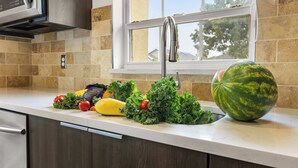 A kitchen with a large window, a stainless steel sink, and various fresh vegetables on the counter.