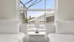 A bedroom with two beds, a small bedside table, and a window with a view of a swimming pool and buildings.