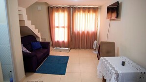 A living room with a sofa, a table with a lace tablecloth, a fan, and a TV mounted on the wall. - Casa Pet Friendly com Varanda e Churrasqueira (Rio das Ostras)