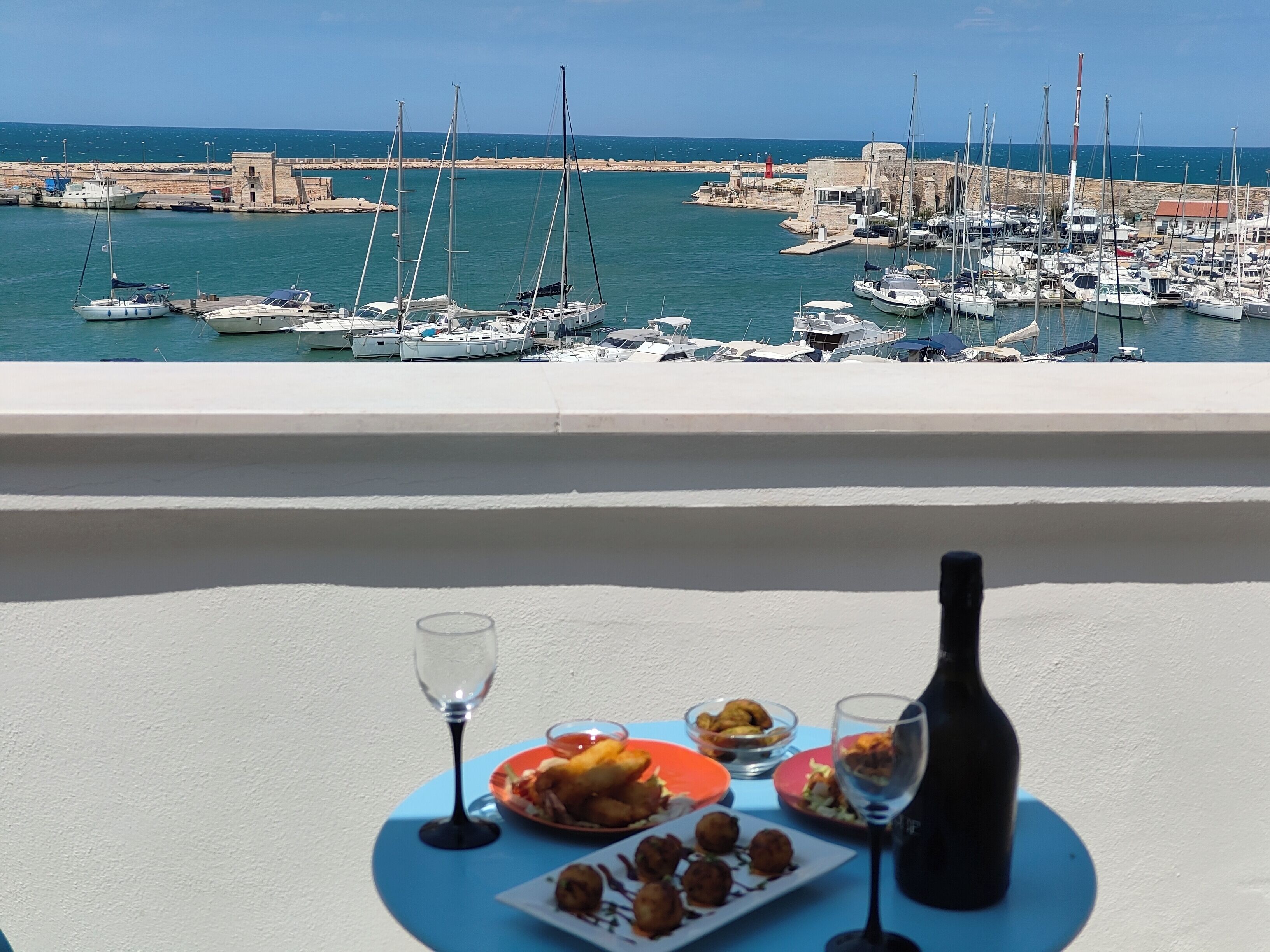 A balcony with a table set for a meal, overlooking a marina with boats and a historic stone wall.