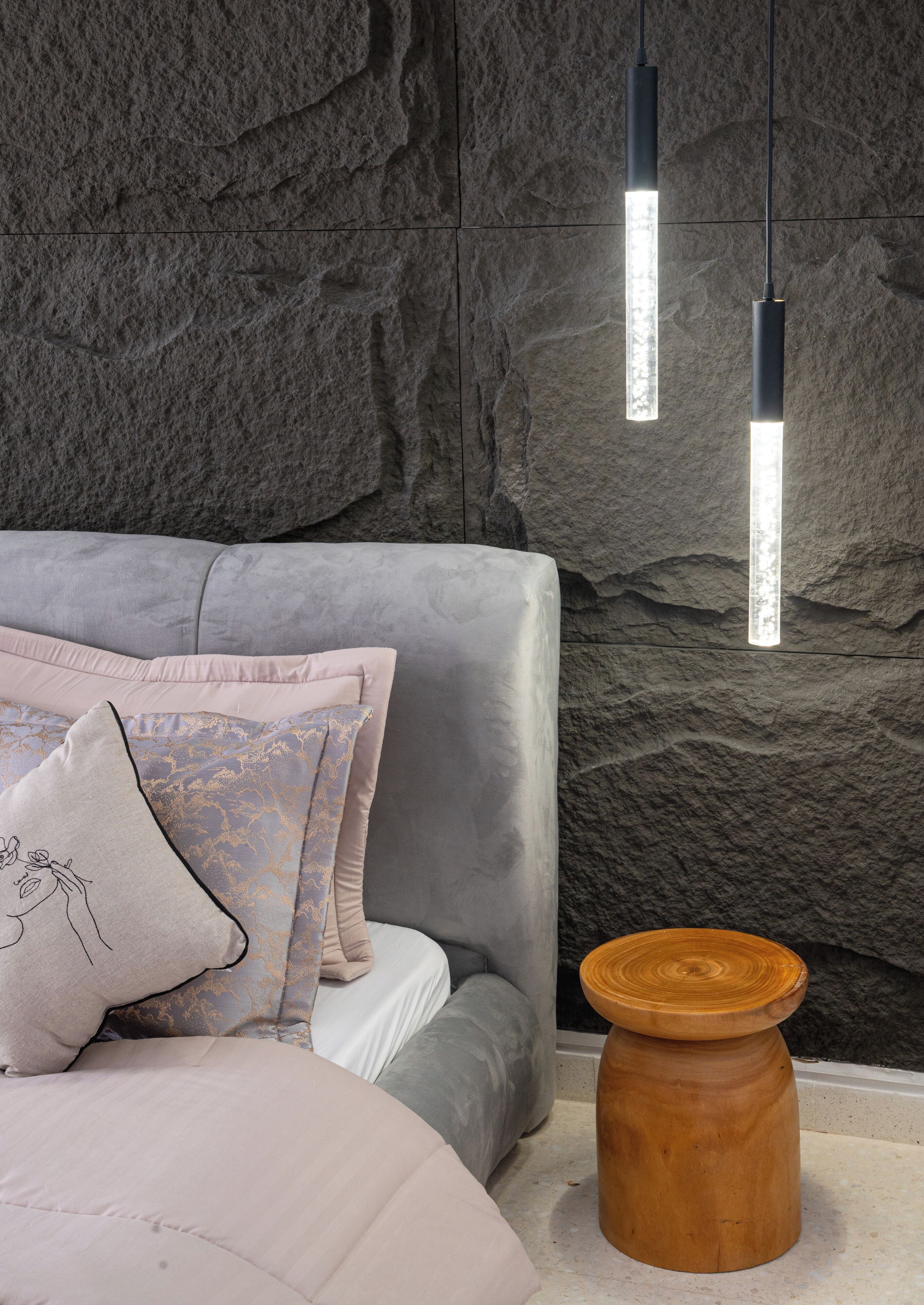 A modern bedroom with a grey upholstered headboard, a wooden side table, and a textured stone wall.