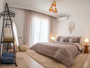 Um quarto com cama, luminárias de cabeceira, luminária pendente, ar-condicionado na parede, um sonho apanhado e um varal.