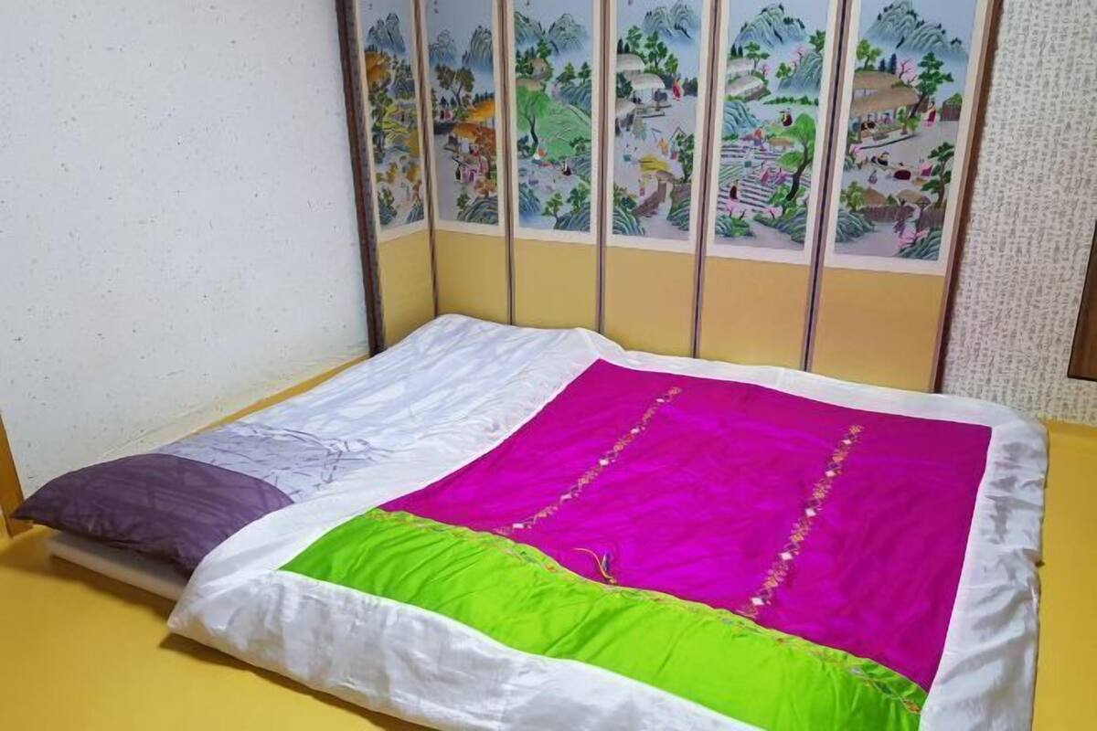 A bed with a white and purple pillow, a green and pink blanket, and a wall with vertical panels.