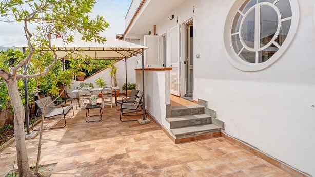 Eine Terrasse mit Gartenmöbeln, einem Baum und einem Gebäude mit rundem Fenster und Treppe.