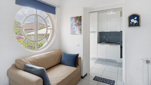 Ein Zimmer mit einem beigefarbenen Sofa, einem runden Fenster, einer Küchenzeile und einer Tür zu einem weiteren Raum.
