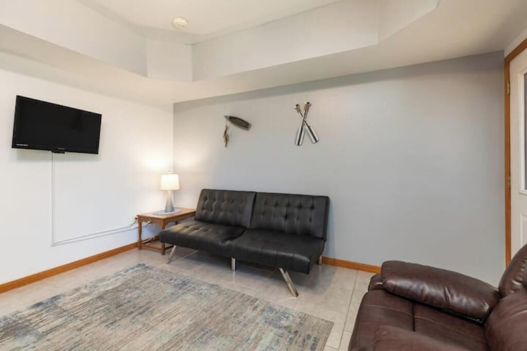 A living room with a black leather sofa, a wooden side table with a lamp, a flat-screen TV mounted on the wall, and a large rug on the floor.