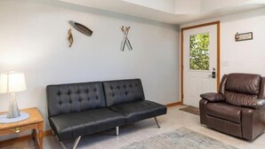 Un salon avec un canapé noir, un fauteuil inclinable en cuir marron, une table d’appoint en bois avec une lampe, et un tableau encadré accroché au mur.