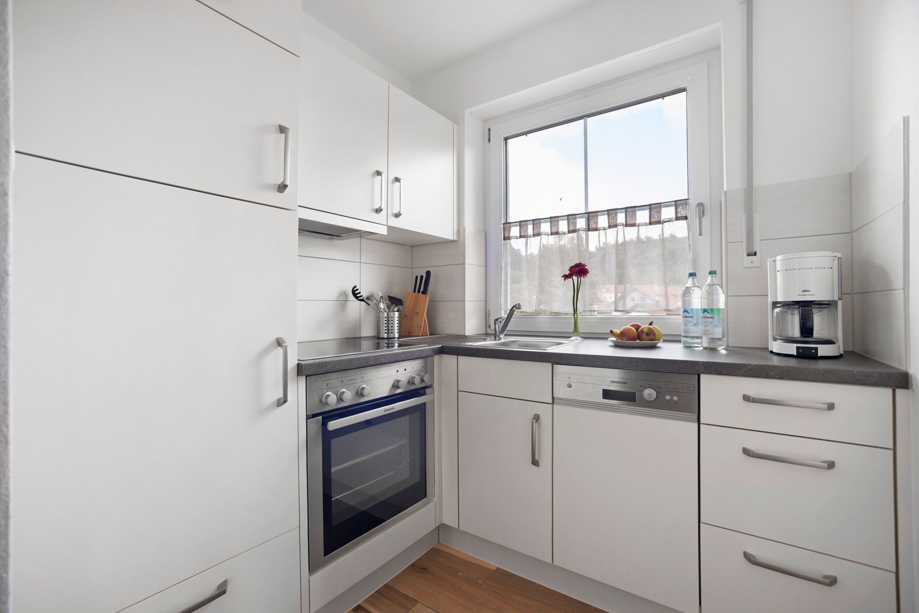 A modern kitchen with white cabinets, a built-in oven, a dishwasher, and a window with a view.