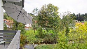 A balcony with a large umbrella, a wooden bench, and a view of a garden with a stream and trees. - Holiday Apartment 'Am Mühlbach' with Shared Garden, Balcony and Wi-Fi (Marktoberdorf)