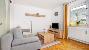 A modern living room with a grey sofa, wooden coffee table, TV stand, and a window with curtains. - Holiday Apartment 'Am Mühlbach' with Shared Garden, Balcony and Wi-Fi (Marktoberdorf)