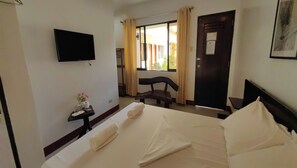 Habitación de hotel con cama, televisor montado en la pared, mesita con artículos de aseo, una silla y ventana con cortinas.