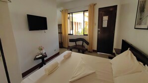 Quarto de hotel com uma cama, televisão presa na parede, uma mesinha com produtos de higiene pessoal, uma cadeira e uma janela com cortinas.