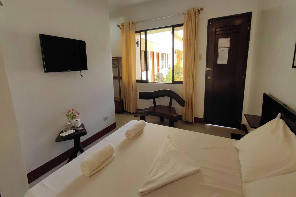 A hotel room with a bed, a TV mounted on the wall, a small table with toiletries, a chair, and a window with curtains.