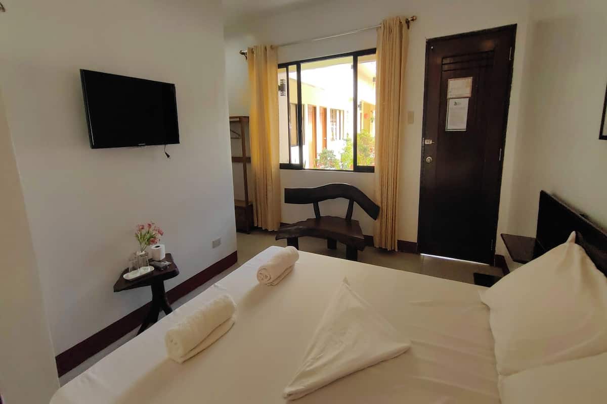 A hotel room with a bed, a TV mounted on the wall, a small table with toiletries, a chair, and a window with curtains.