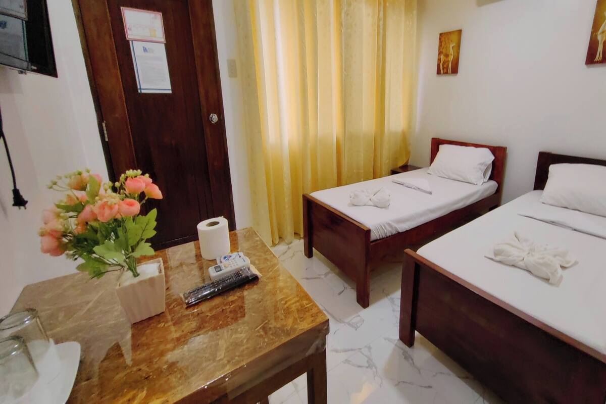 A hotel room with two single beds, a wooden table with a vase of flowers, a remote control, and a roll of toilet paper.