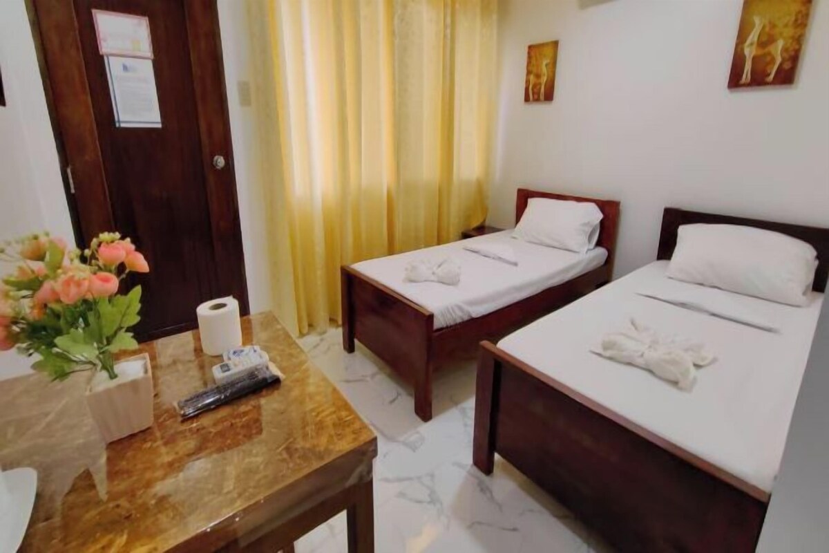 A hotel room with two beds, a wooden table with a vase of flowers, a remote control, and a roll of toilet paper.