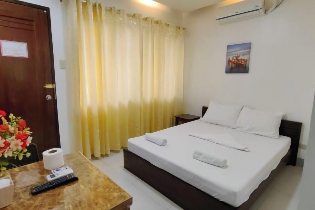 A hotel room with a bed, a bedside table with a remote control, a roll of toilet paper, and a vase with flowers.