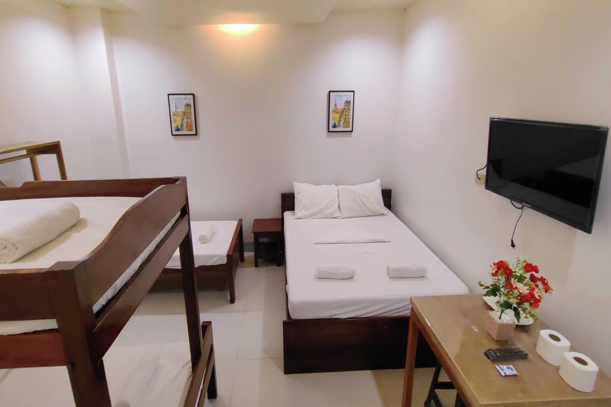 A hotel room with a bed, a desk with a TV, a chair, a small table with a vase of flowers, and two framed pictures on the wall.
