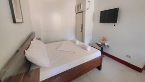 A neatly made bed with white linens and a rolled towel, a small bedside table with a vase of flowers, a wall-mounted TV, and a door with a mirror. - Pring Pension House Puerto Princesa By Zuzu (Puerto Princesa)