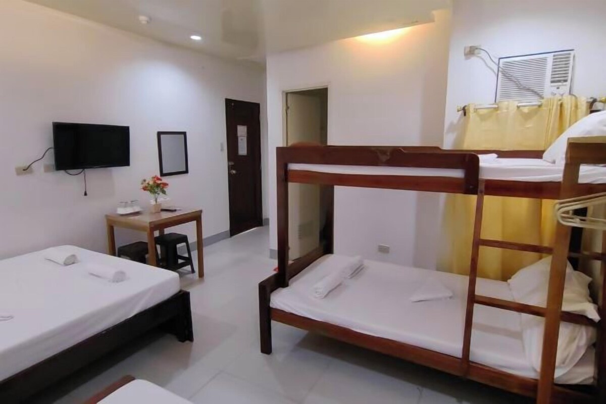 A hotel room with a bunk bed, a single bed, a desk with a chair, a TV mounted on the wall, and a window with a curtain.