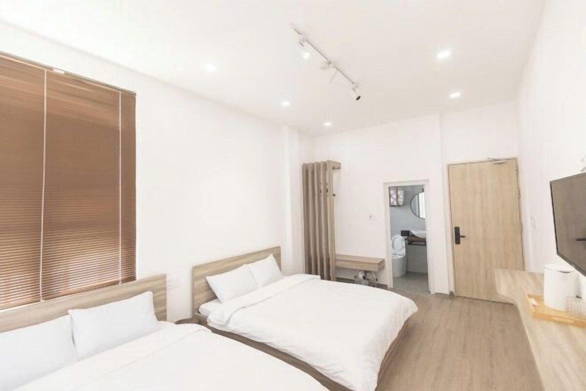 A modern hotel room with two beds, a wooden floor, a TV, and a bathroom visible through an open door.