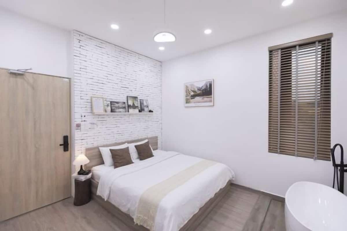 Ein Schlafzimmer mit einem Bett, einem Nachttisch, einem Wandbild, einem Fenster mit Jalousien und einem Badezimmer mit Waschbecken.