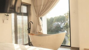 Ein modernes Badezimmer mit einer freistehenden Badewanne, einem großen Fenster mit Jalousien und einem an der Wand befestigten Flachbildfernseher.