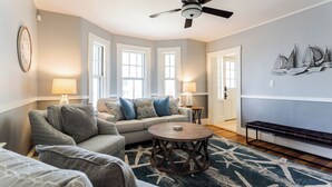 Un salon avec un canapé, une table basse ronde et un ventilateur de plafond. Deux lampes sont posées sur des tables d’appoint, et un banc est placé contre le mur.