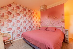 A bedroom with a patterned wallpaper, a bed with pink bedding, a wicker chair, a bedside table, and a hanging light fixture.