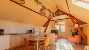 Een ruime keuken met witte kasten, een eethoek met een ronde tafel en oranje stoelen, en een slaapverdieping met een roze fauteuil.