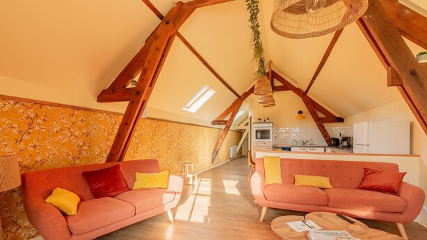 Een knus loft-achtig vertrek met een hoge plafond, houten balken, een zithoek, een kleine tafel en een keuken in de achtergrond.