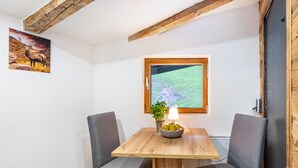 A dining area with a wooden table, two grey chairs, a bowl of fruit, a potted plant, and a window with a view of a green landscape. - Room '1 - Cozy' with Wi-Fi (Fieberbrunn)