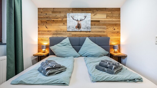 A double bed with two pillows, a wooden headboard, a framed deer picture, bedside lamps, and folded towels. - Room '1 - Cozy' with Wi-Fi (Fieberbrunn)