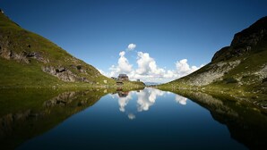 A serene lake with a house in the distance, surrounded by grassy hills.