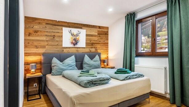 A bedroom with a bed, wooden panel wall, artwork, window with curtains, and a nightstand with a lamp.