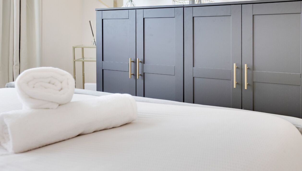 A neatly made bed with a rolled white towel, a dark grey wardrobe with gold handles, and a small table with a glass top and a candle.