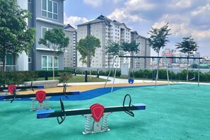 An outdoor exercise area with seesaws and a blue rubber surface, surrounded by greenery and buildings. - 121 Residences by manatidur (Petaling Jaya)