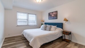 A bedroom with a bed, a nightstand, a lamp, a window with blinds, and a painting on the wall.